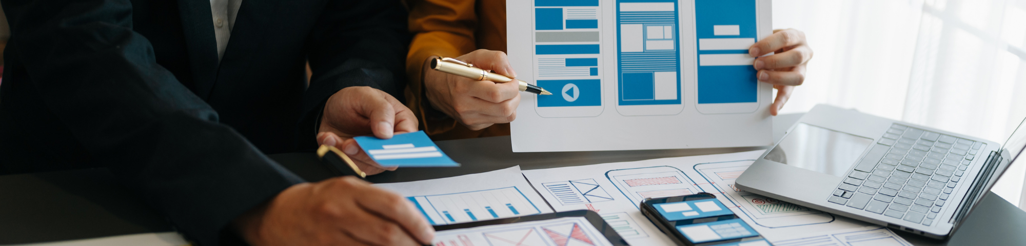 Designers collaborating. The image shows hands pointing at and discussing different mobile and tablet layout designs using printed sheets and a tablet."