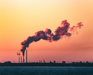 Industrial smokestacks emitting large clouds of smoke against a vibrant sunset sky, symbolizing pollution and environmental impact.